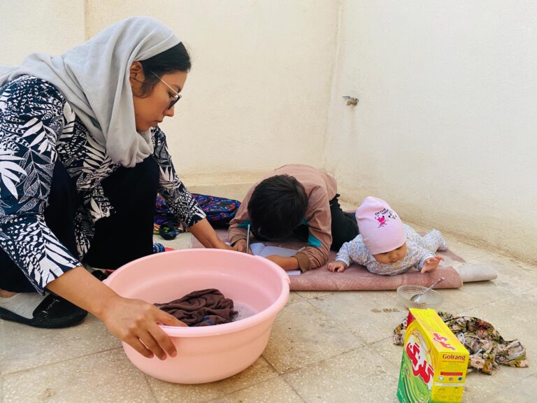 Hazara woman from Afghanistan shelters inside a house in Iran, washing clothes while her children study and play. Credit Atefa Mohseni, 2024